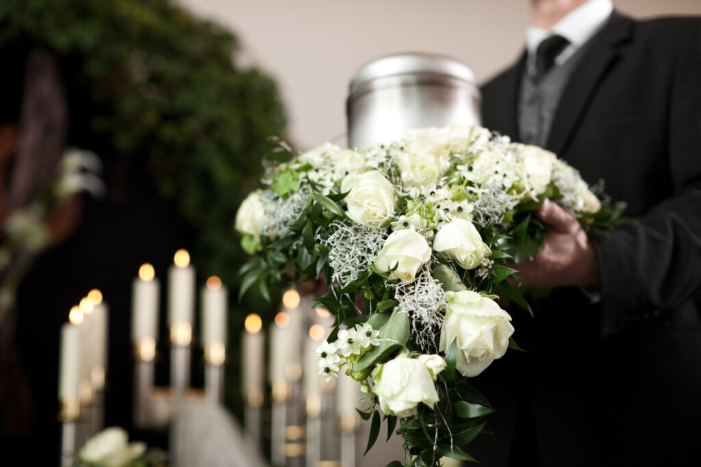 Ein Mann hält eine silberne Urne und einen weißen Blumenkranz bei einer Trauerfeier zur Feuerbestattung.